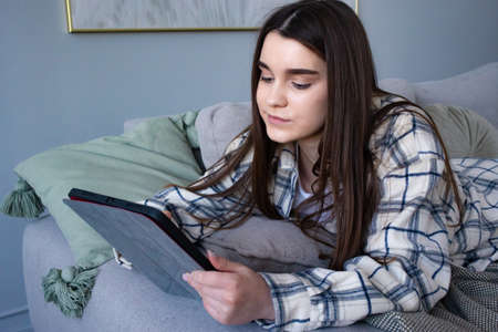 Caucasian young woman looks at the tablet lying on couch in modern home and using wireless internet....
