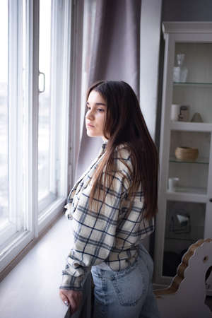 Thoughtful brown haired female student in casual outfit looking away while standing near window at...