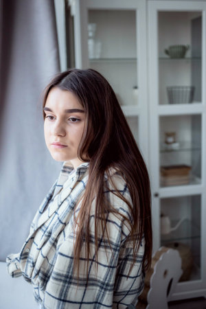 Thoughtful brown haired female student in casual outfit looking away while standing near window at homeの写真素材