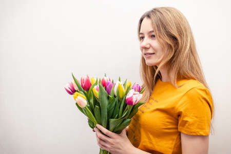 attractive girl in an orange t-shirt holding a bunch of colorful tulips in hands on a light...