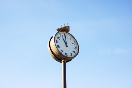 A quartz clock mounted on a metal pole displays the time as 1155 against a vibrant blue sky...