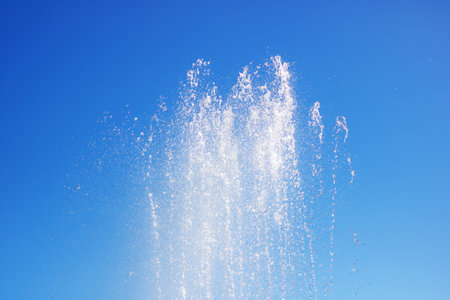 A liquid fountain spraying water against an electric blue sky, creating a stunning natural landscape with trees and clouds in the backgroundの写真素材