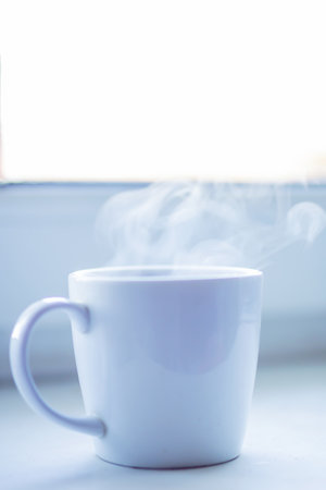 A traditional cup of coffee is resting on a sunny window sill, with steam rising gracefully from its surface, creating a cozy atmosphereの写真素材