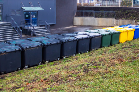 A neat row of trash cans is strategically lined up in front of a large building, serving their...