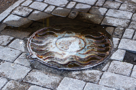A heavy manhole cover is resting on a cobblestone sidewalk that is located beside a large puddle of...