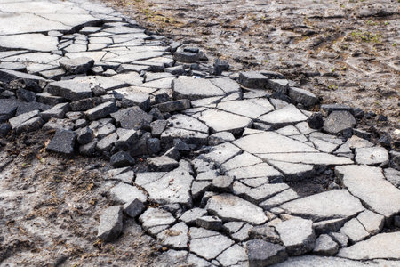 This image features a closeup view of a cracked asphalt road, illustrating the wear and tear over...