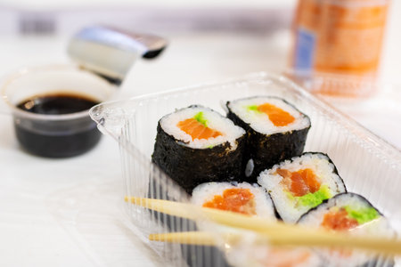 A vibrant plastic container filled to the brim with delicious sushi rolls and two elegant chopsticks resting beside itの写真素材