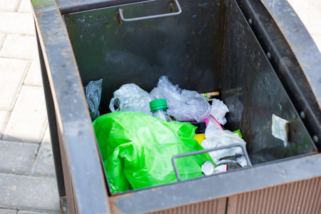A trash can that is overflowing and filled with an abundance of plastic bags and bottles, indicating...