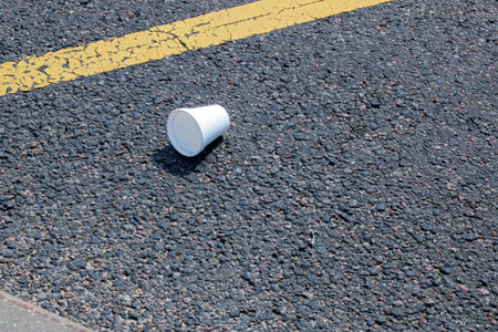 A discarded white disposable cup lying on a rough asphalt surface, highlighting environmental...