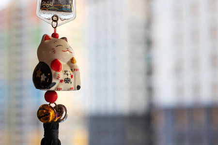lucky cat charm hanging near window tiny ceramic figurine with bell, red beads and black tassel, warm morning sunlightの写真素材