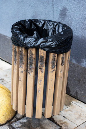 weathered wooden slat trashcan, black liner, stained slats and grimy exterior against concrete wall on damp sidewalkの写真素材