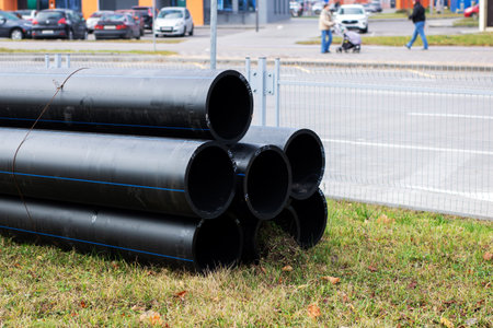 Stacked black pipes on roadside grass, large pvc conduits arranged in neat pyramid, parked cars and...
