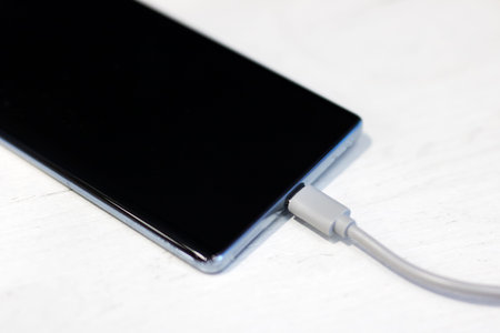 Smartphone connected to charging cable, lying on light wood surface, closeup of usb connector,...