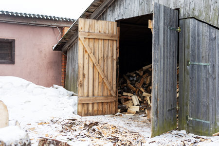 Open woodshed revealing stacked firewood and damp floor, snowbanks outside and weathered boards,...