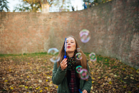 Attractive young woman is enjoying making a bubbles in the public park. Ground covered with autumn leaves.の写真素材