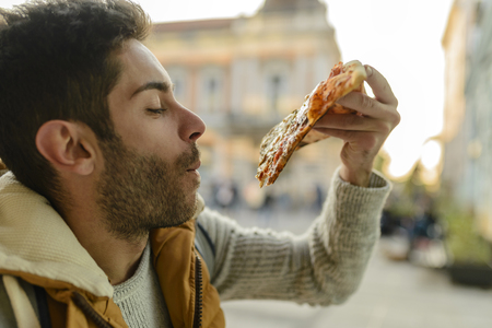 Handsome bearded young man with orange jacket eating pizza in the city streetの写真素材