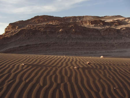 Sand drawn by the wind, with some little rocks showing long shadows caused by afternoon light and a stonewall behind it, in Valle de la Luna, Atacama Desert, Chile.の写真素材