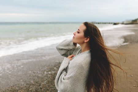 A beautiful young girl walks along the seashore, a storm, hair fly apart, a gray cardigan, a sports figure, in sneakers, enjoys, autumn or winterの写真素材