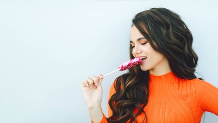 Young beautiful girl with dark curly hair eats a pink lollipop candy screaming proud in a bright...