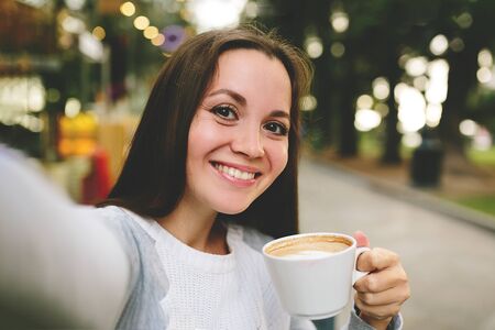 Young beautiful girl with a coffe taking and makes selfie in street cafe.の写真素材