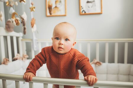 Beautiful little girl in a crib in a cozy light room in a sweater of fashionable shades. learns to stand at the support. cheerful little girl standing in the crib at homeの写真素材