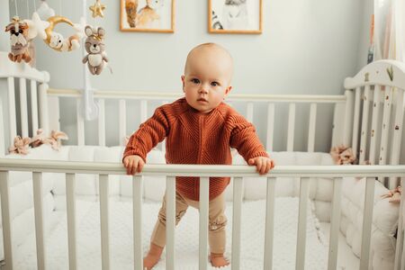 Beautiful little girl in a crib in a cozy light room in a sweater of fashionable shades. learns to stand at the support. cheerful little girl standing in the crib at homeの写真素材