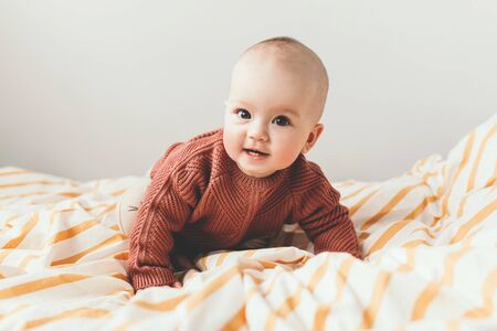 Beautiful little baby girl on the bed in a cozy brown sweater smiles. Concept of motherhood and...