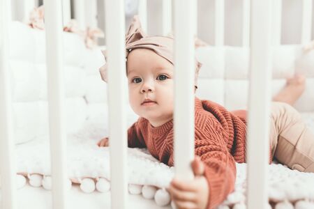 Beautiful little girl in a crib in a cozy light room in a sweater of fashionable shades. learns to...