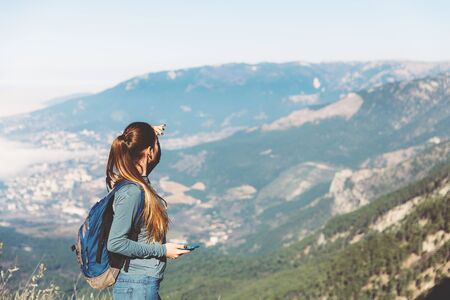 Young beautiful girl travels alone in the mountains in spring or autumn, looks into the distance with phone and enjoys nature, rocks and green forests, view of the landscape. a backpack behind and sportswear, freedom and lightnessの写真素材
