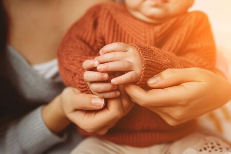 Young mother holding the handles of her little daughter, close-up, little fingers, concept of motherhood, cozy sweater. Mother with her newborn baby care handsの写真素材