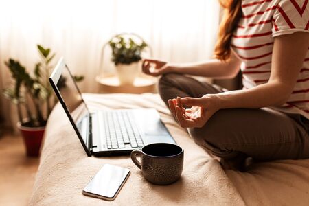 Young calm woman sits in staff easy lotus pose near laptop at home interior, meditating alone on the floor with eyes closed, online yoga training, doing breathing exercise in the morning, no stressの写真素材