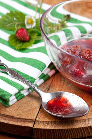 Silver spoon lying on a wooden tabletop with a sample of freshly boiled whole fruit strawberry jam from the glass bowl behindの写真素材