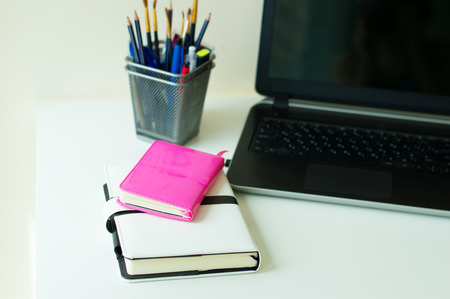 Two notebooks of pink and white color, office stationery and black laptop on white office table