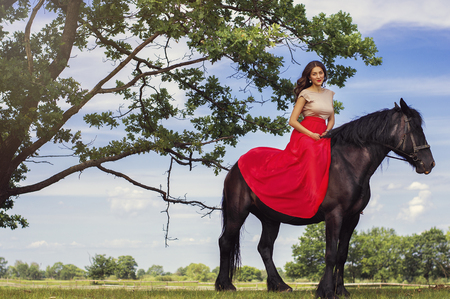 Beautiful woman with trendy make-up and in long red dress sitting on a black horse under a big tree. Horse riding concept.の写真素材