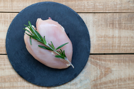 Background with raw chicken breast and fresh green rosemary on black slate plate on old wooden tableの写真素材