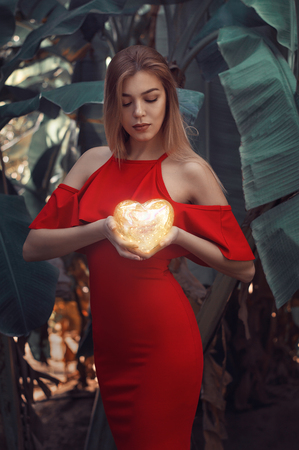 Blond female model in sexy red dress holding in her arms a small shining heart. Woman standing near big banana leaves. Fine artの写真素材