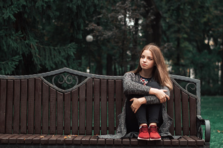 One teenage girl with blond hair in knitted cardigan and black denim jeans sitting on wooden bench...