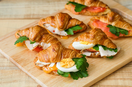 Croissant sandwiches served on wooden board. Croissants with egg, melted cheese, parsley, chicken ham, smoked salmonの写真素材