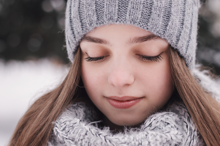 Portrait of female teenager with long hair, in knitted hat and scarf of gray color looking down. Beatiful face of a young girl. Winter time. Warm clothingの写真素材