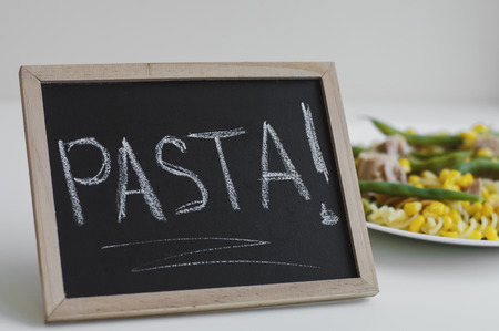 Word "Pasta" written on a small framed blackboard and a portion of fusilli pasta with tuna, corn and green beans on white tableの写真素材