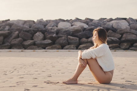 Female model in white sweater and beige bikini sitting on a sandy beach during sunset. Relax, leisure and summer vacationの写真素材