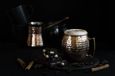 Dark chocolate with nuts, cappuccino coffee in a brass mag served on a black slate board with...