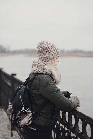 Young female with blond hair dressed in khaki jacket, knitted scarf, hat and with backpack standing...