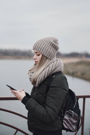 Teenage female with blond hair, backpack and dressed in knitted hat and scarf standing outside and looking at smartphone screen. Online communication, social networks conceptの写真素材