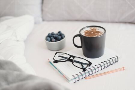 Cappuccino with cinnamon in black mug, note book, pen, reading glasses and blueberries served in bed. Work from bed concept. Studing and workong from homeの写真素材
