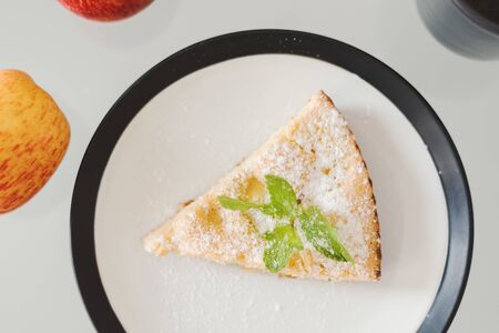 Piece of apple pie with powdered sugar and fresh mint served on a white plate with black borders,...