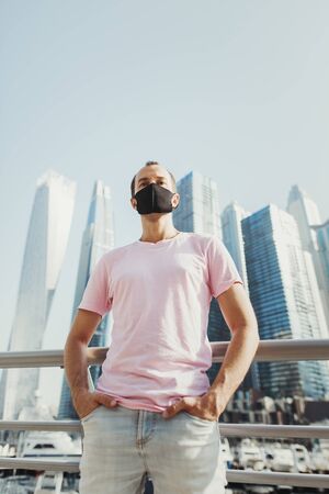 Young man in blue jeans and pink t-shirt wearing protective face mask of black color and standing in city business center with office buildings. Prevention of Covid 19の写真素材
