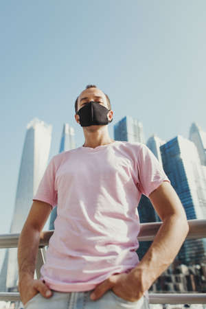 Confident young man in pink t-shirt and jeans wearing protective face mask of black color. Male tourist standing at city business center during Covid 19 pandemicの写真素材