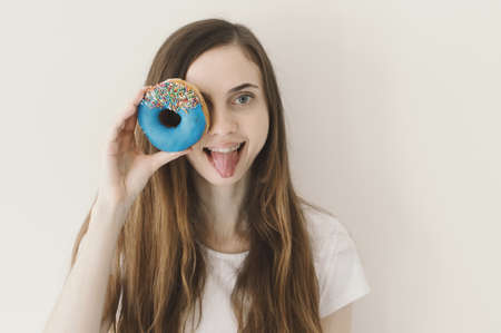 Funny portrait of young European woman holding a blue donut with sprinkles near her eye and showing...
