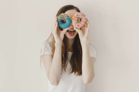Funny portrait of a woman holding blue and pink donuts near her eyes and showing a tongueの写真素材